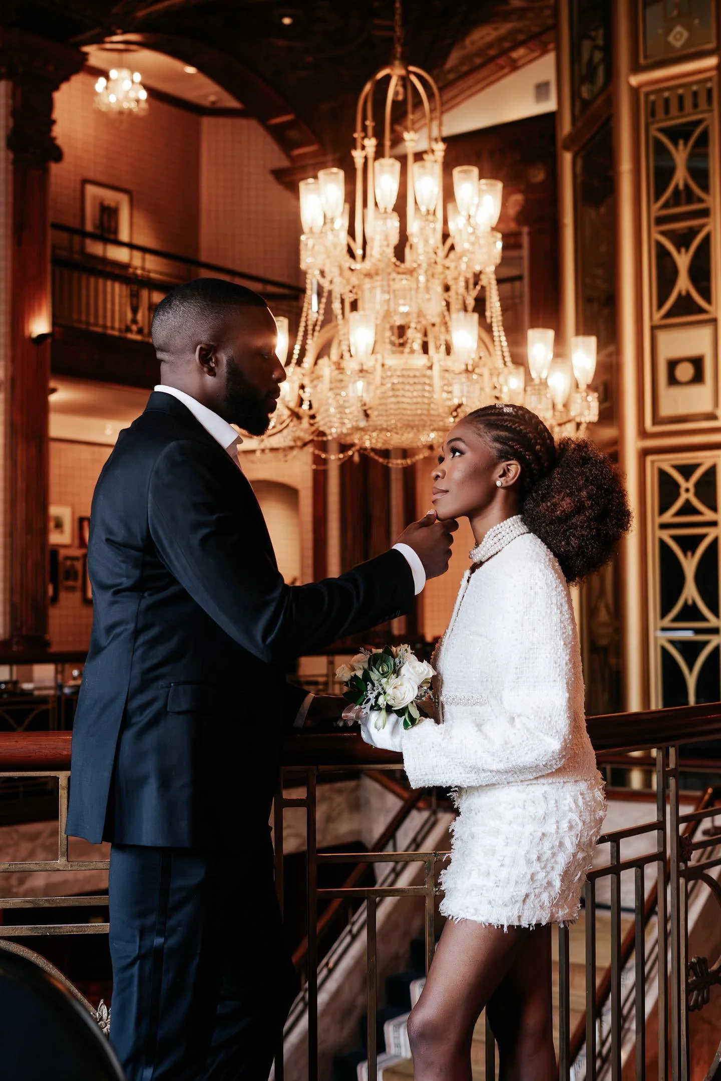 A man in a black suit gently touches a woman's chin as they look into each other's eyes in an elegantly decorated room with a grand chandelier overhead.