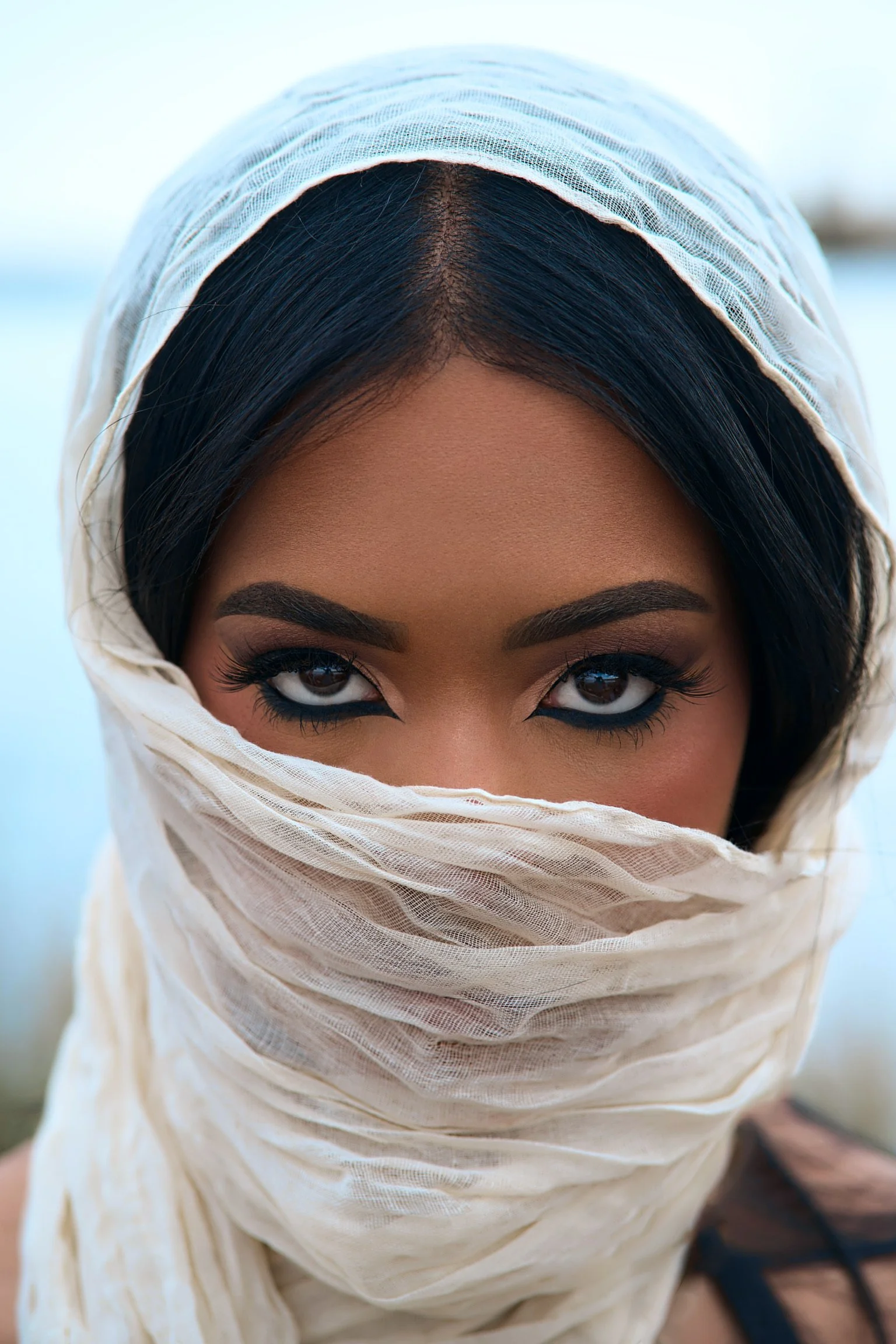 Close-up of a woman with dark hair and makeup, mostly covering her face with a light beige headscarf, only revealing her intense brown eyes.