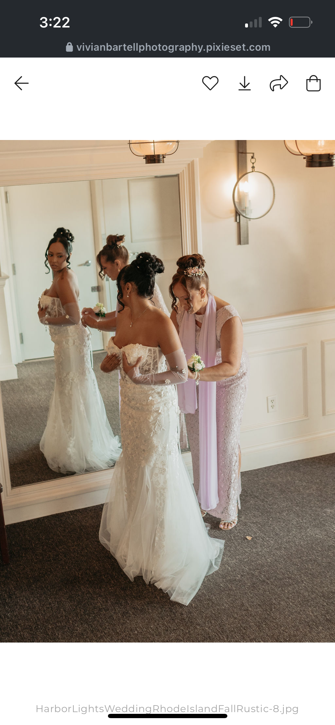 A bride in a lace wedding gown is getting ready with help from an older woman, likely her mother, in a pink dress, while her reflection is visible in a large mirror. The room has warm lighting and elegant fixtures.
