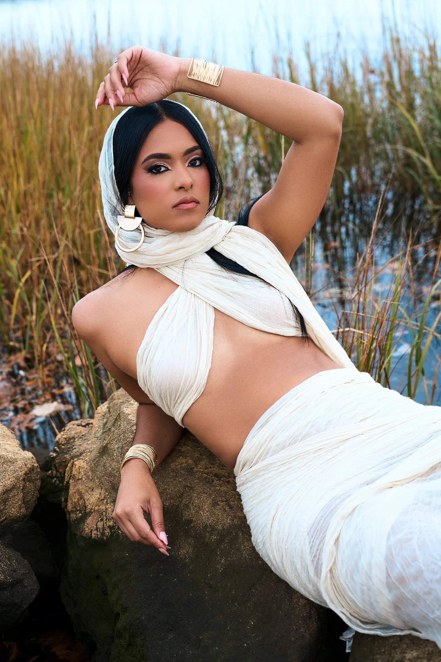 A woman with dark hair and makeup, wearing a cream-colored headscarf, earrings, and accessories, posed outdoors near water and tall grass, reclining on rocks.