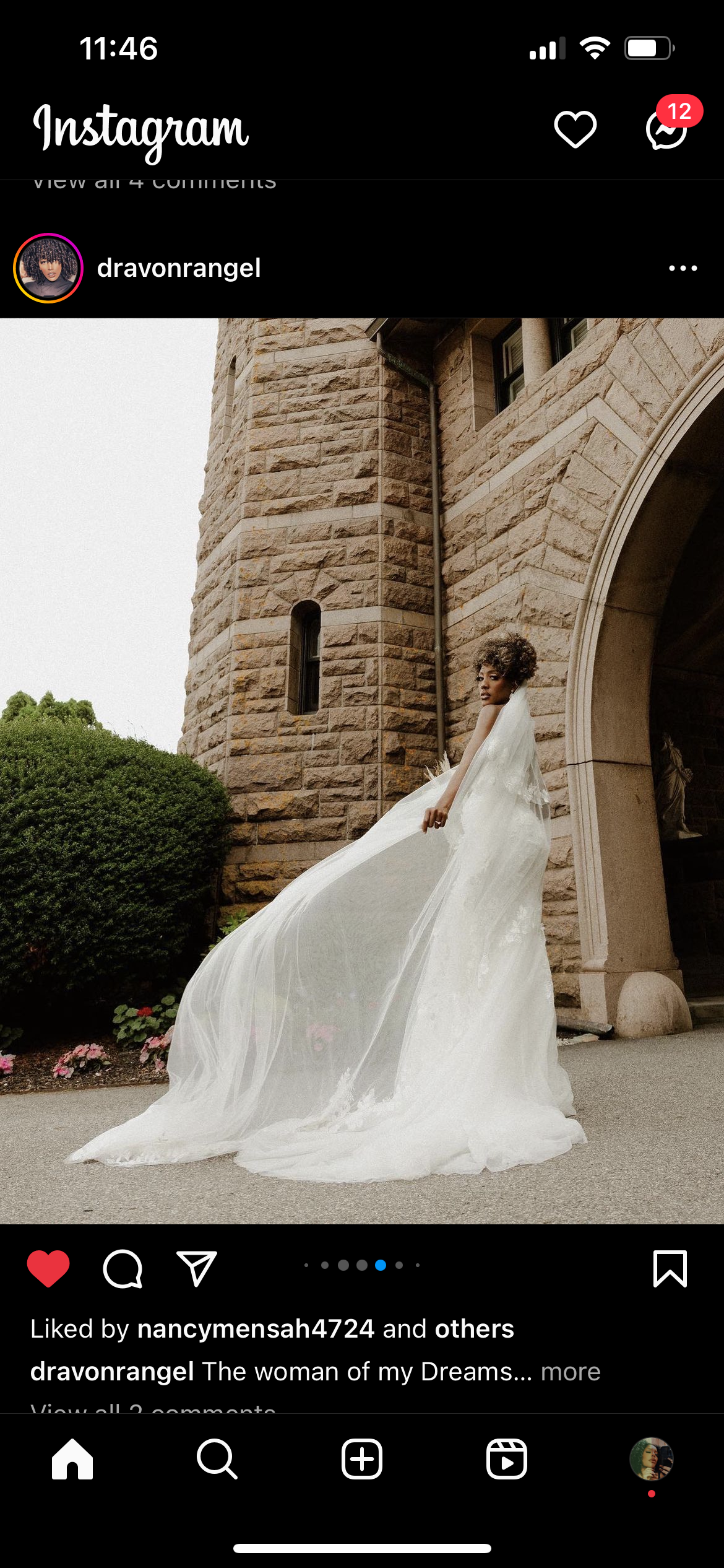 A woman in a white wedding gown standing outside a stone building with a columned archway, garden bushes, and pink flowers nearby.