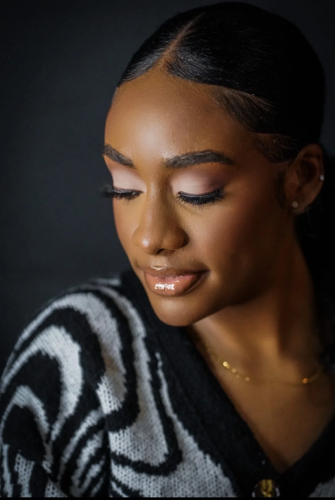 Close-up portrait of a young woman with smooth skin, dark hair slicked back, wearing a black and white patterned sweater, with closed eyes and glossy lips, against a dark background.