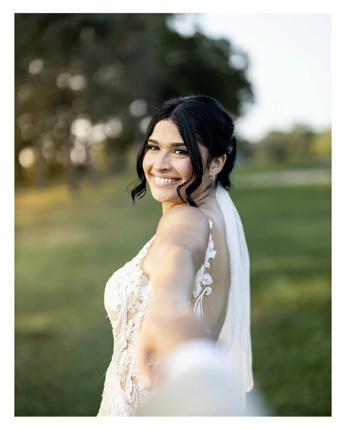 A smiling woman in a white lace dress outdoors, holding the camera with her outstretched arm, with a blurred green park background.
