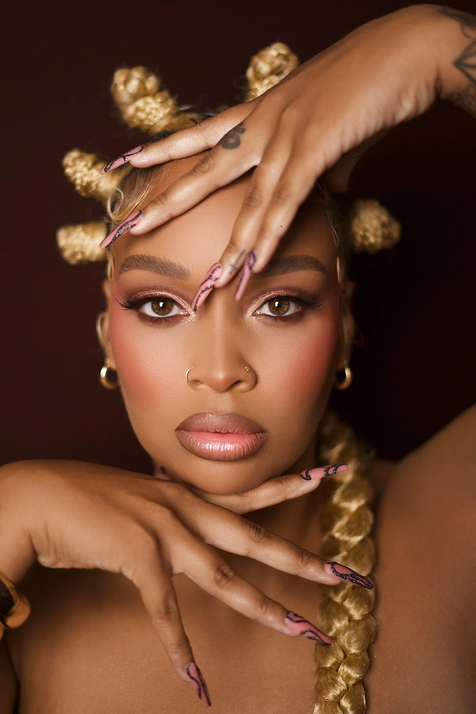 A close-up portrait of a woman with blonde hair styled in bantu knots, wearing makeup with shimmering eye shadow, and gold hoop earrings. She is posing with her hands framing her face against a dark background.