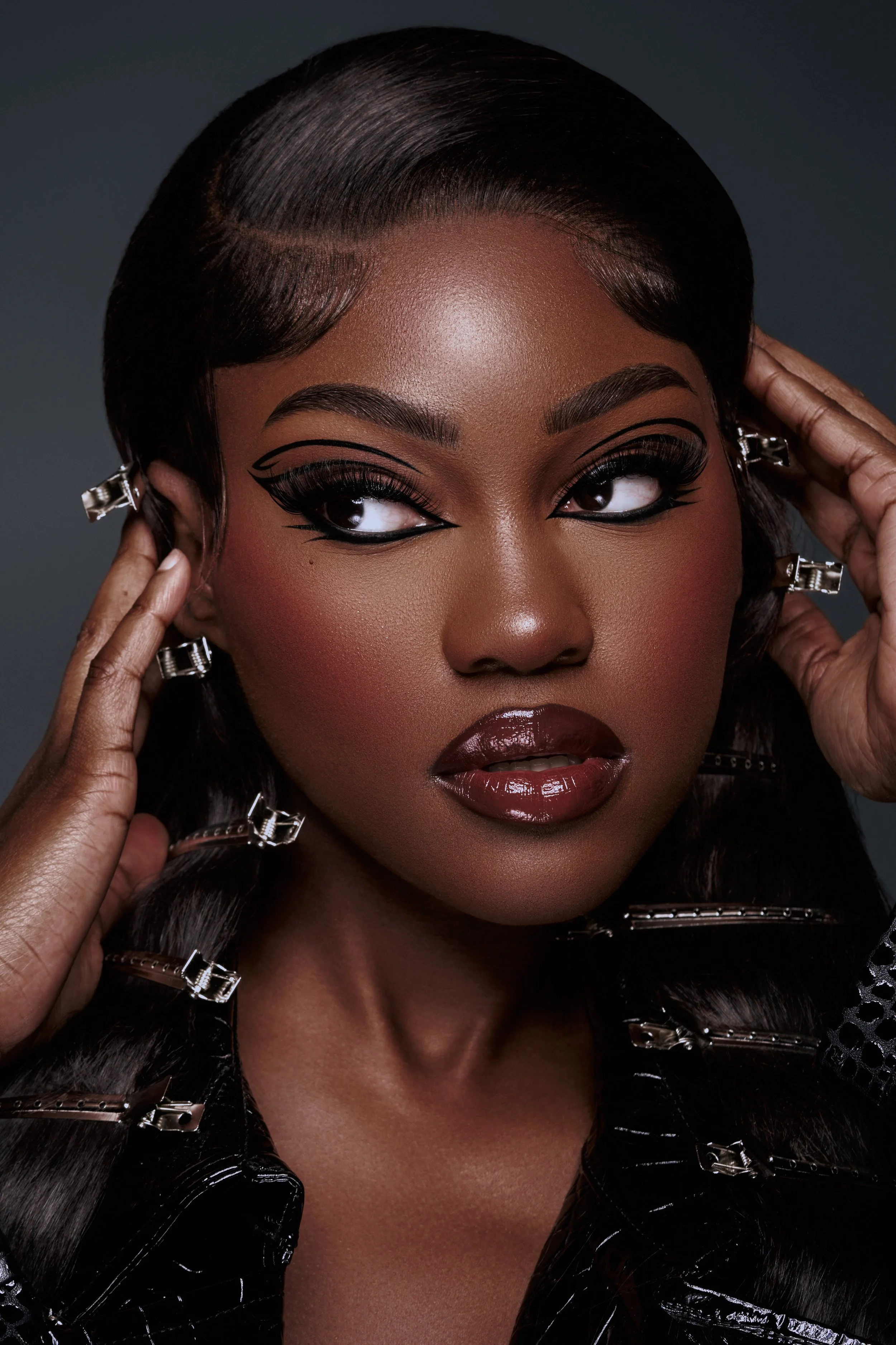 Close-up of a woman with dark skin, styled hair, bold makeup, and large hoop earrings, holding her hair with both hands.