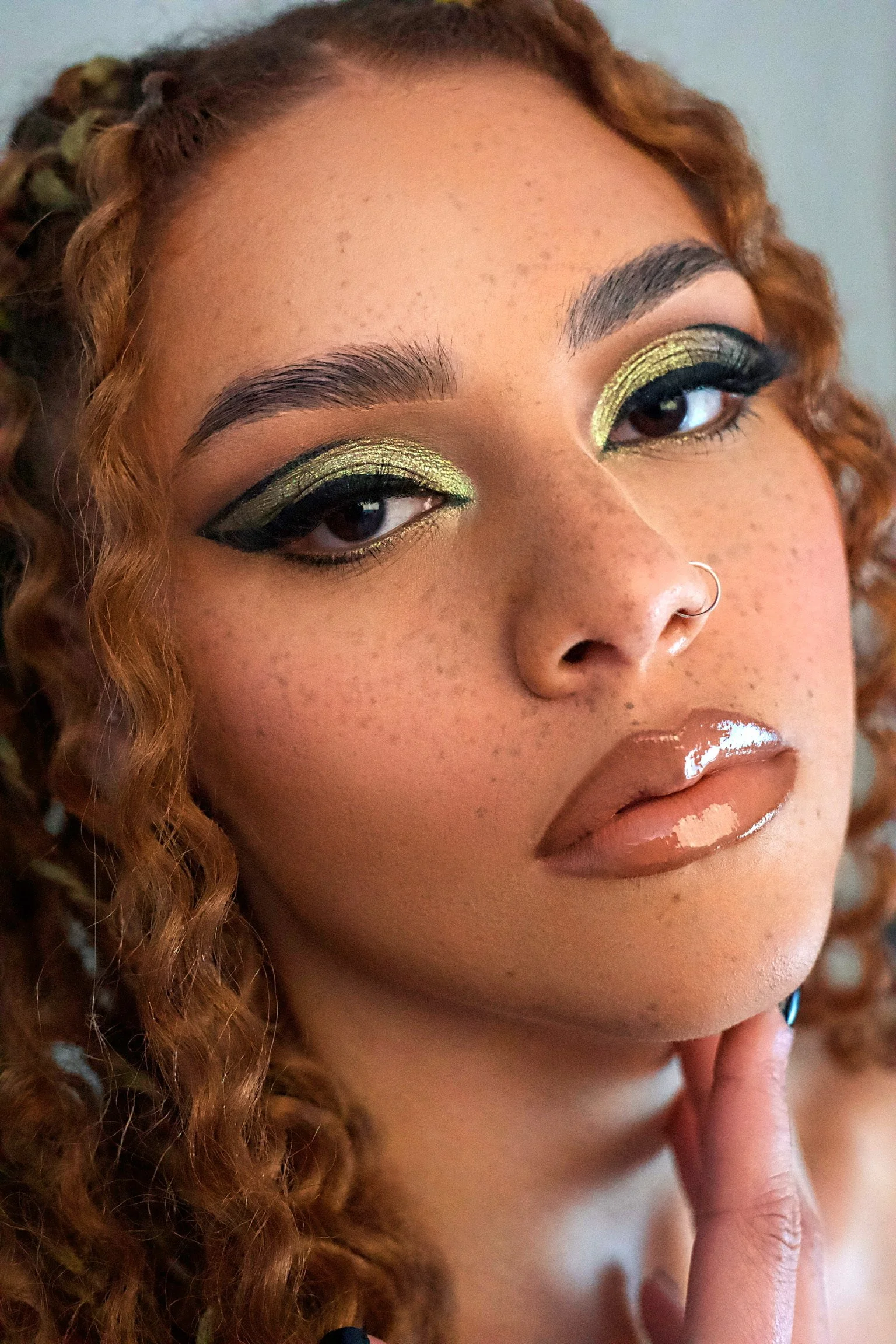 Close-up of a woman with curly hair, gold and black eye makeup, a nose ring, and glossy nude lipstick.