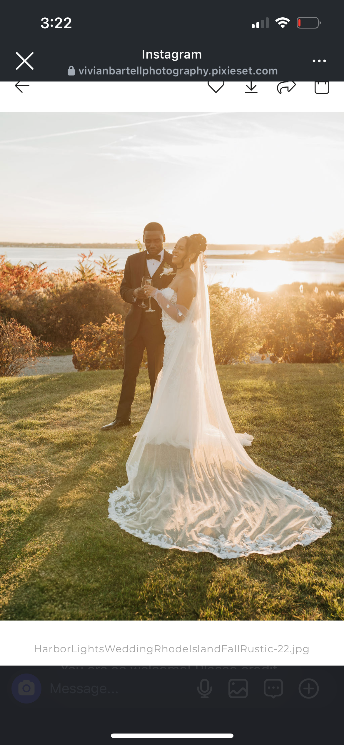 A bride and groom in wedding attire standing outside near a lake during sunset, with the bride smiling while holding a glass, and the groom looking at the bride, surrounded by fall foliage.
