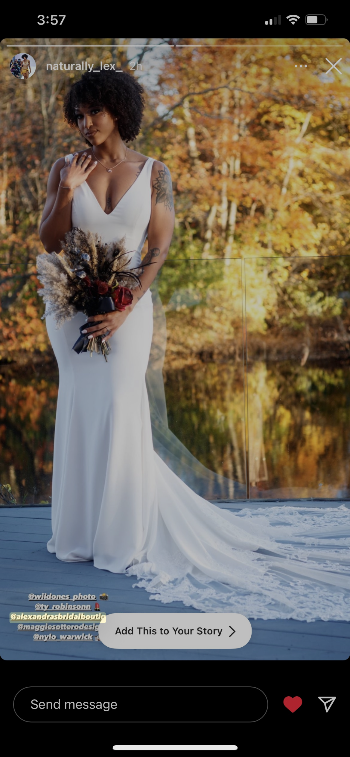 A woman in a white wedding dress holding a bouquet of dried flowers, standing outdoors with autumn trees and a body of water in the background.