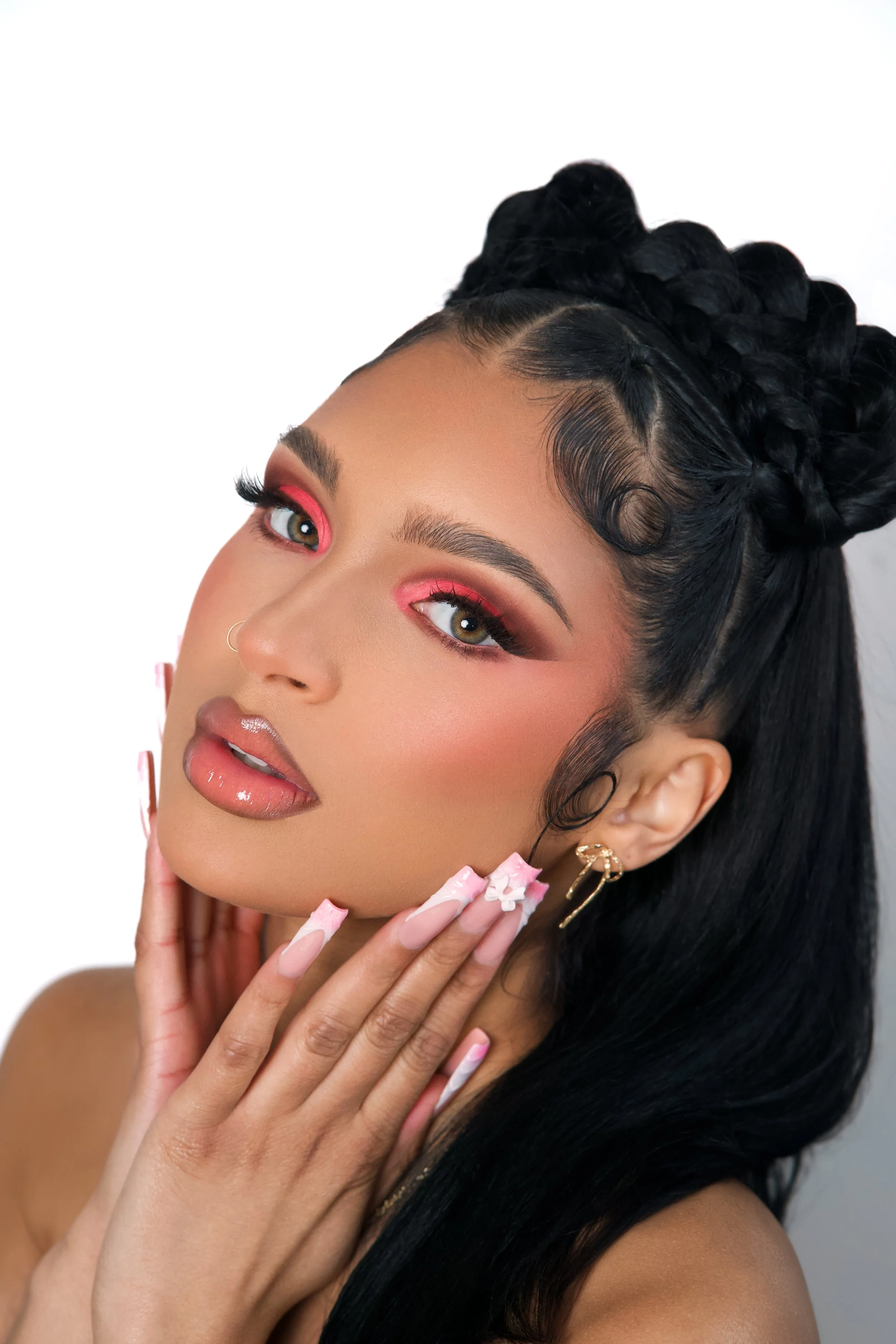 A woman with makeup, styled black hair in braids with curls, and wearing gold earrings, touching her face with manicured nails.