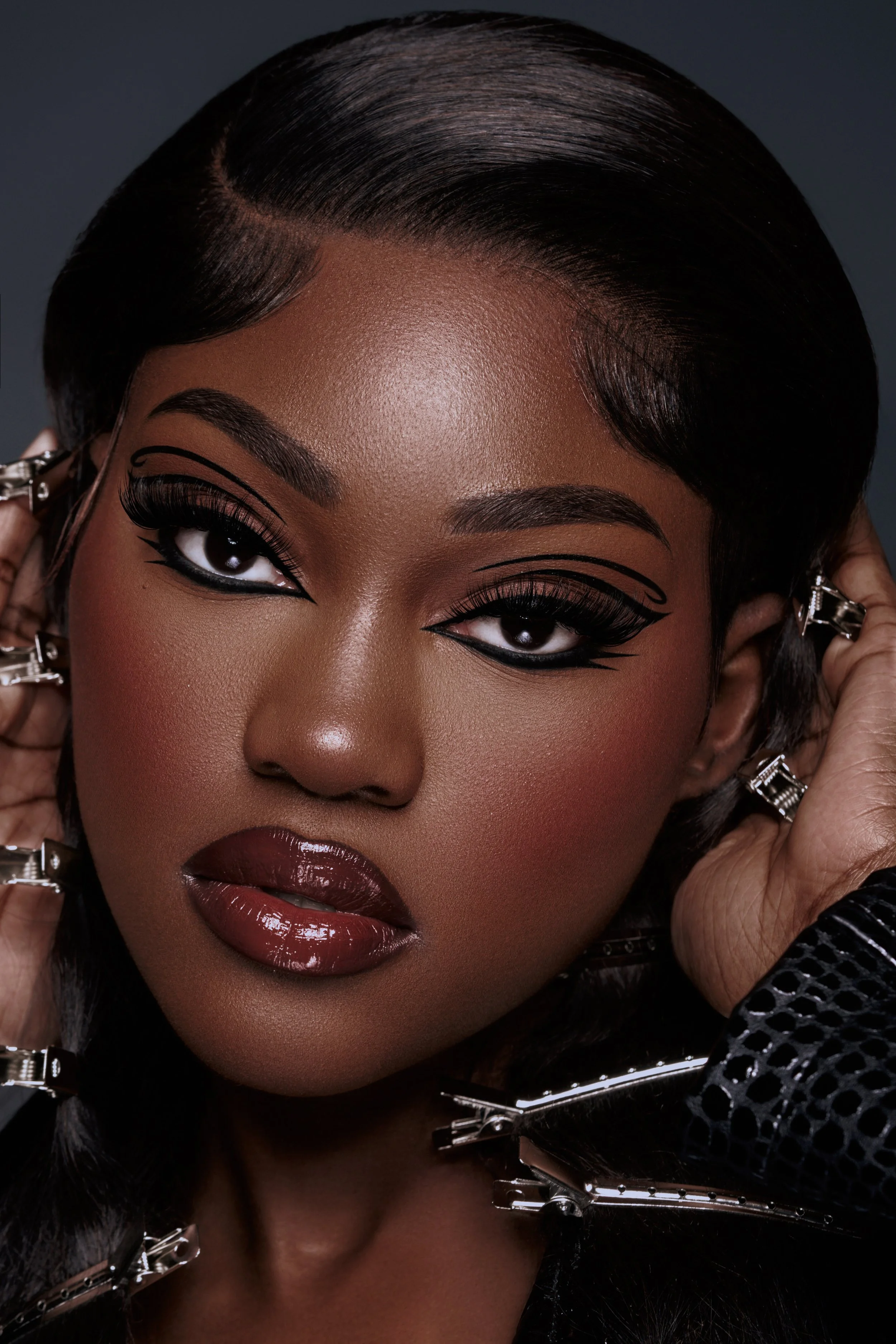 Close-up portrait of a woman with short black hair, makeup, and jewelry, holding clip pins in her hair.