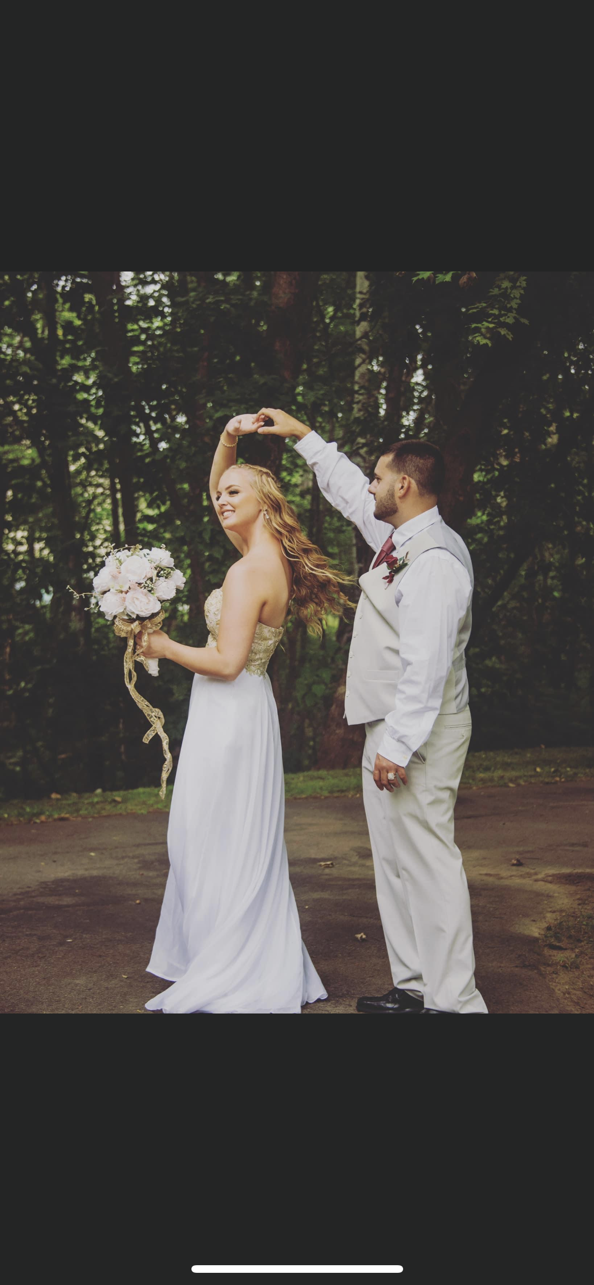 A newlywed couple dance outdoors in a forest setting, with the groom twirling the bride who holds a bouquet of flowers.