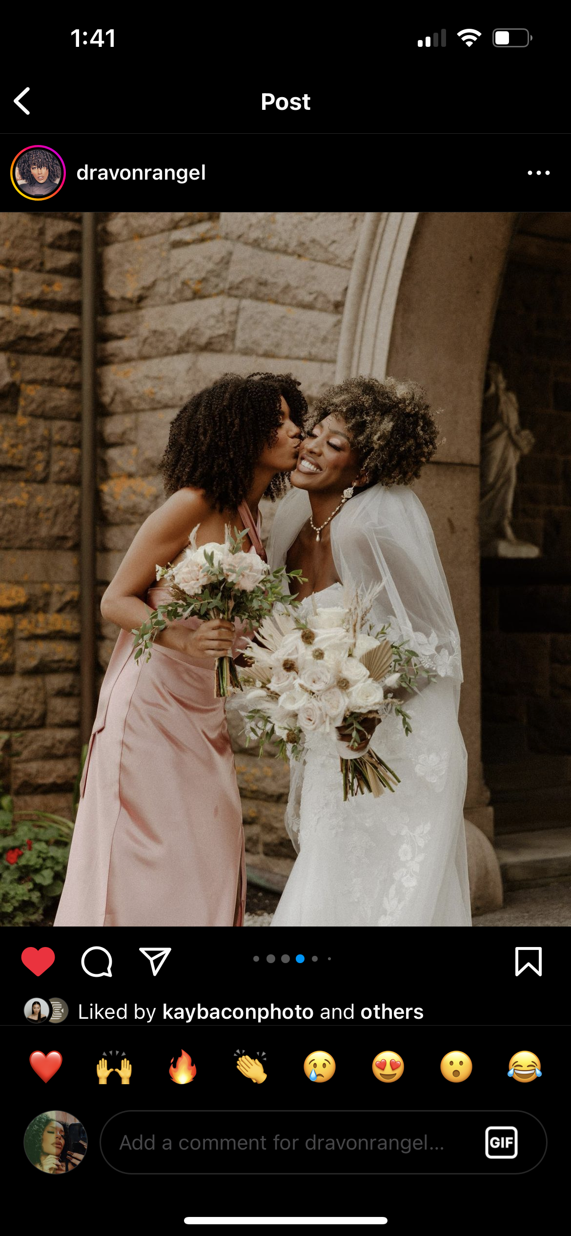 Two women sharing a loving moment, one dressed in a wedding gown and the other in a pink dress, each holding a bouquet of flowers, standing outside a stone building.
