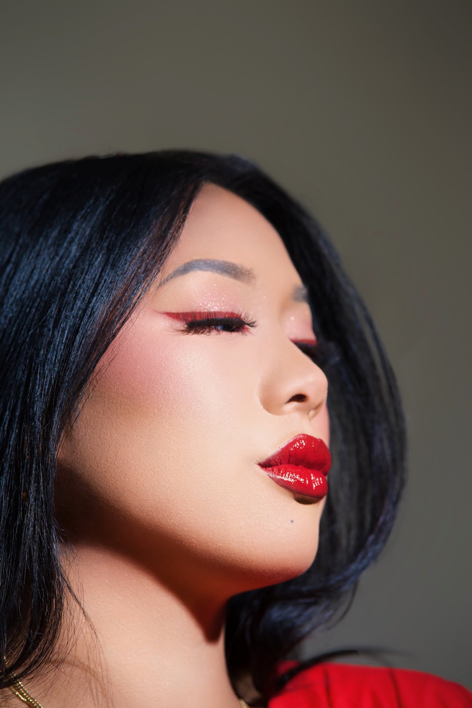 Close-up of a woman with dark, wavy hair and red makeup, including red lipstick and eyeliner, with her eyes closed.