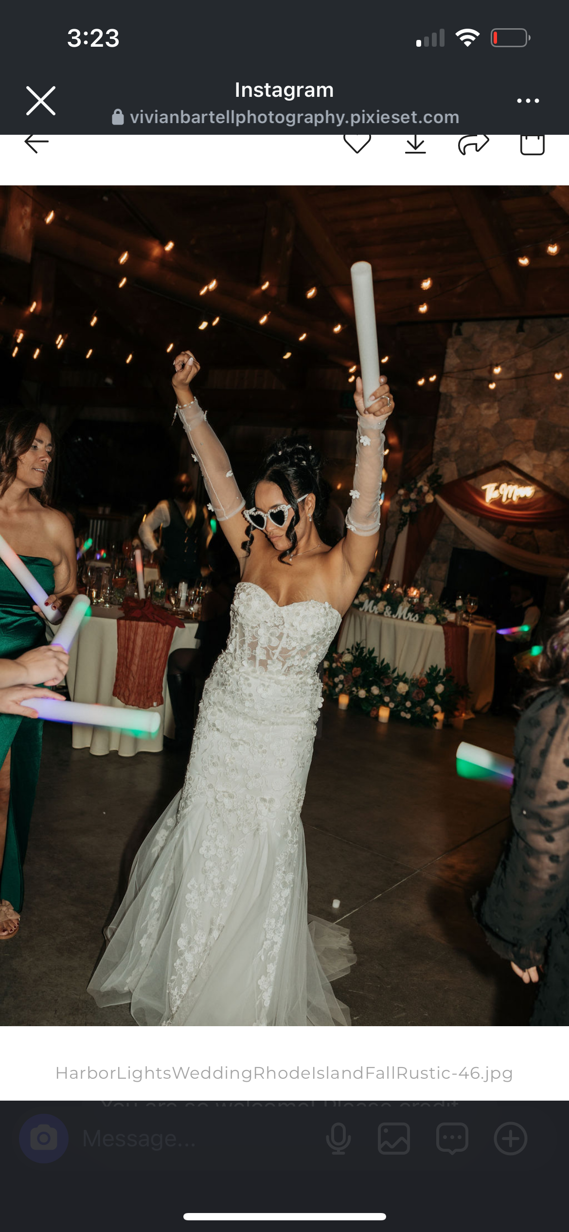 Bride dancing with glow sticks at her wedding reception, wearing a white lace wedding gown and heart-shaped sunglasses, surrounded by guests in a decorated rustic venue with string lights and floral arrangements.