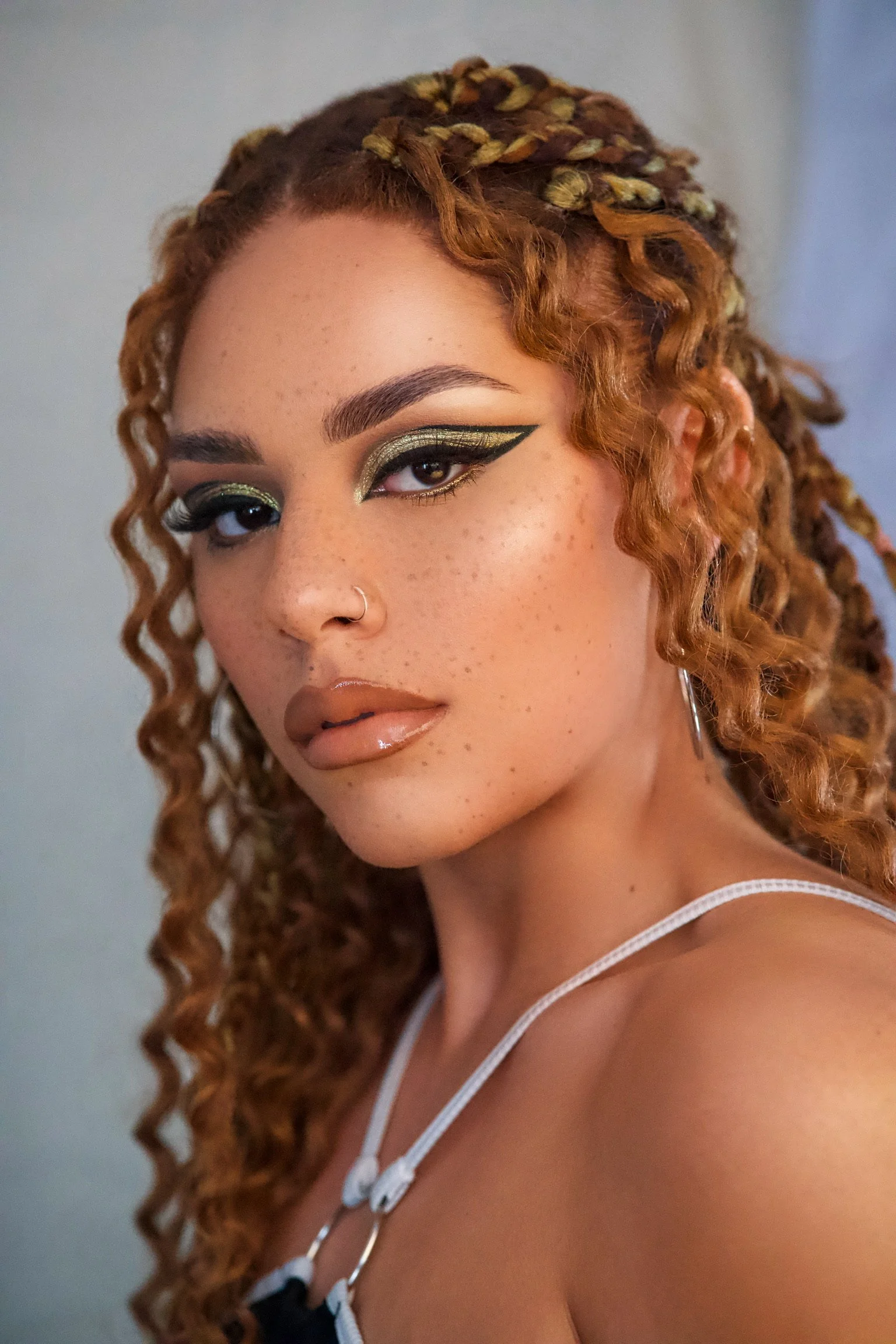 A young woman with curly reddish hair styled in braids, wearing bold eyeliner and shimmering green eyeshadow, with nude lipstick, a nose ring, and hoop earrings.