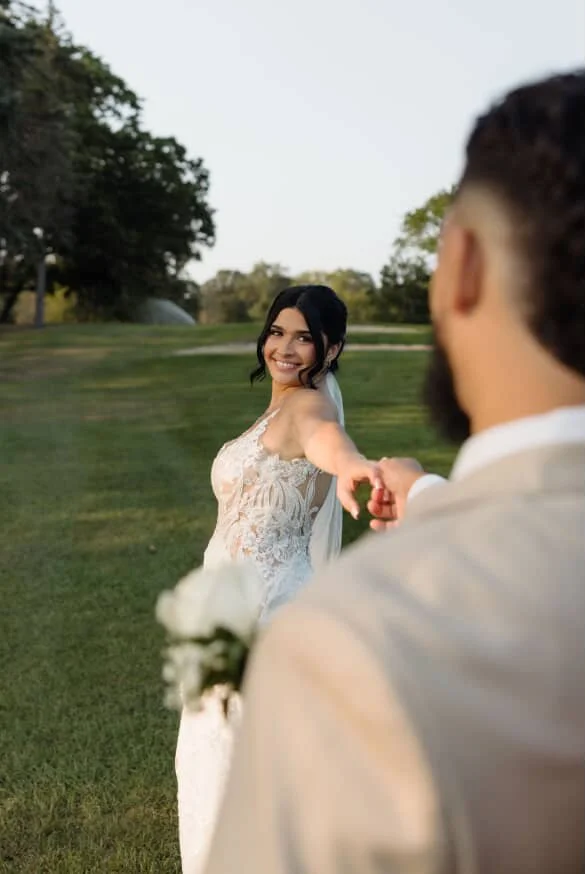 A bride wearing a lace wedding dress smiling and holding hands with a groom, who is seen from behind, in an outdoor park setting.