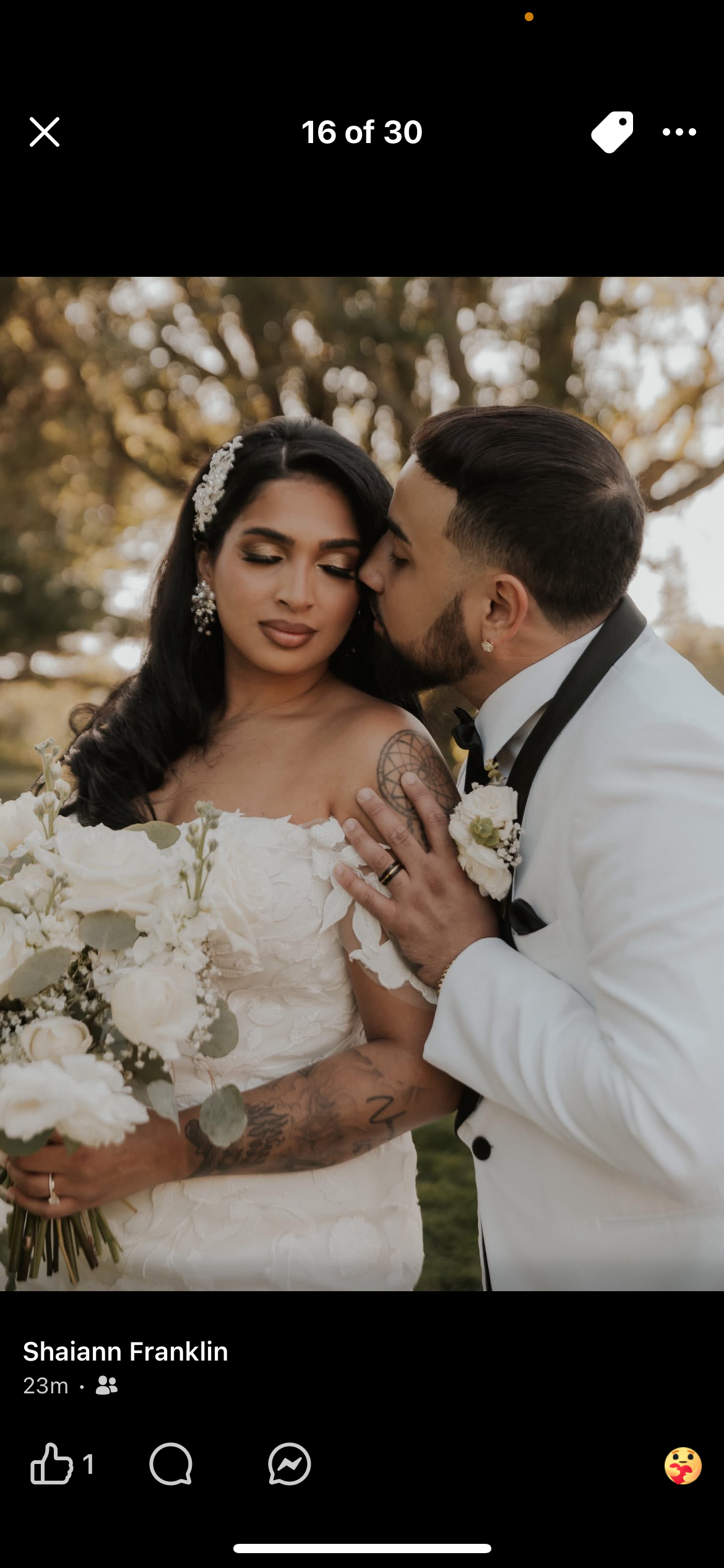 A bride and groom are close together outdoors during a wedding. The groom is kissing the bride's temple while she has her eyes closed. The bride holds a bouquet of white flowers, and she has tattoos on her arms and shoulders. The groom is wearing a w