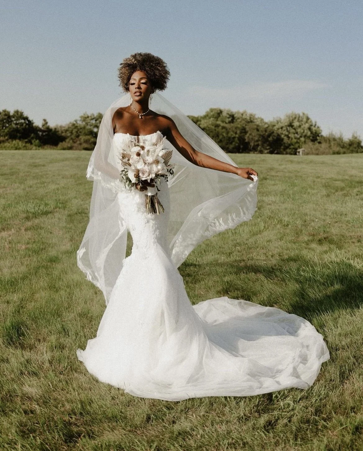 A woman in a strapless white wedding dress holding a bouquet, standing on a grassy field with clear blue sky in the background.