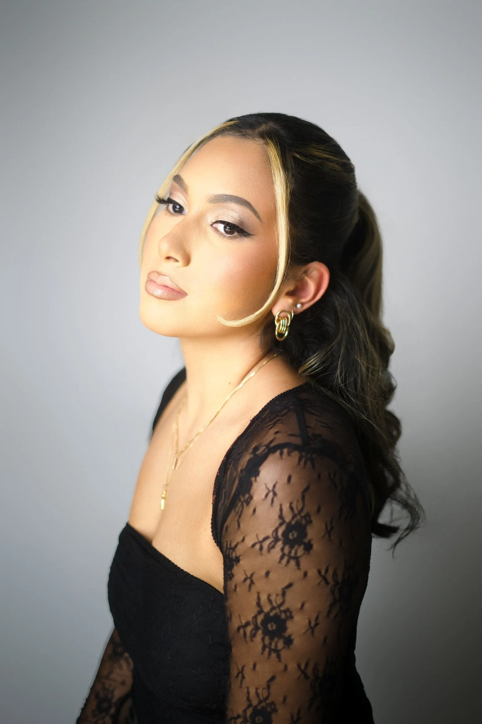 A woman with long dark and blonde hair, wearing a black lace top, gold jewelry, and makeup, looks confidently at the camera against a plain gray background.