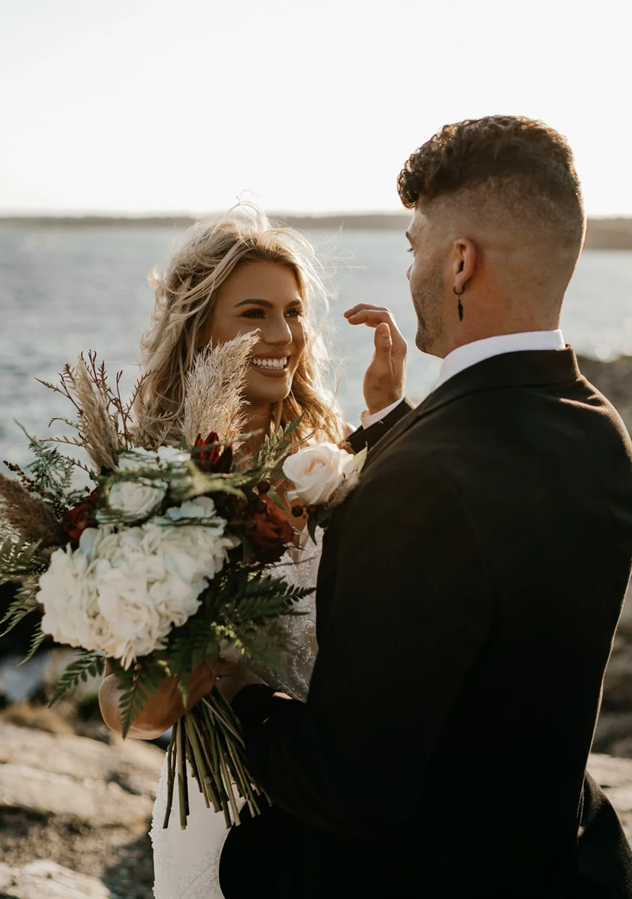 A bride and groom exchanging vows outdoors near a body of water during sunset, with the bride holding a bouquet of flowers and both dressed in wedding attire.