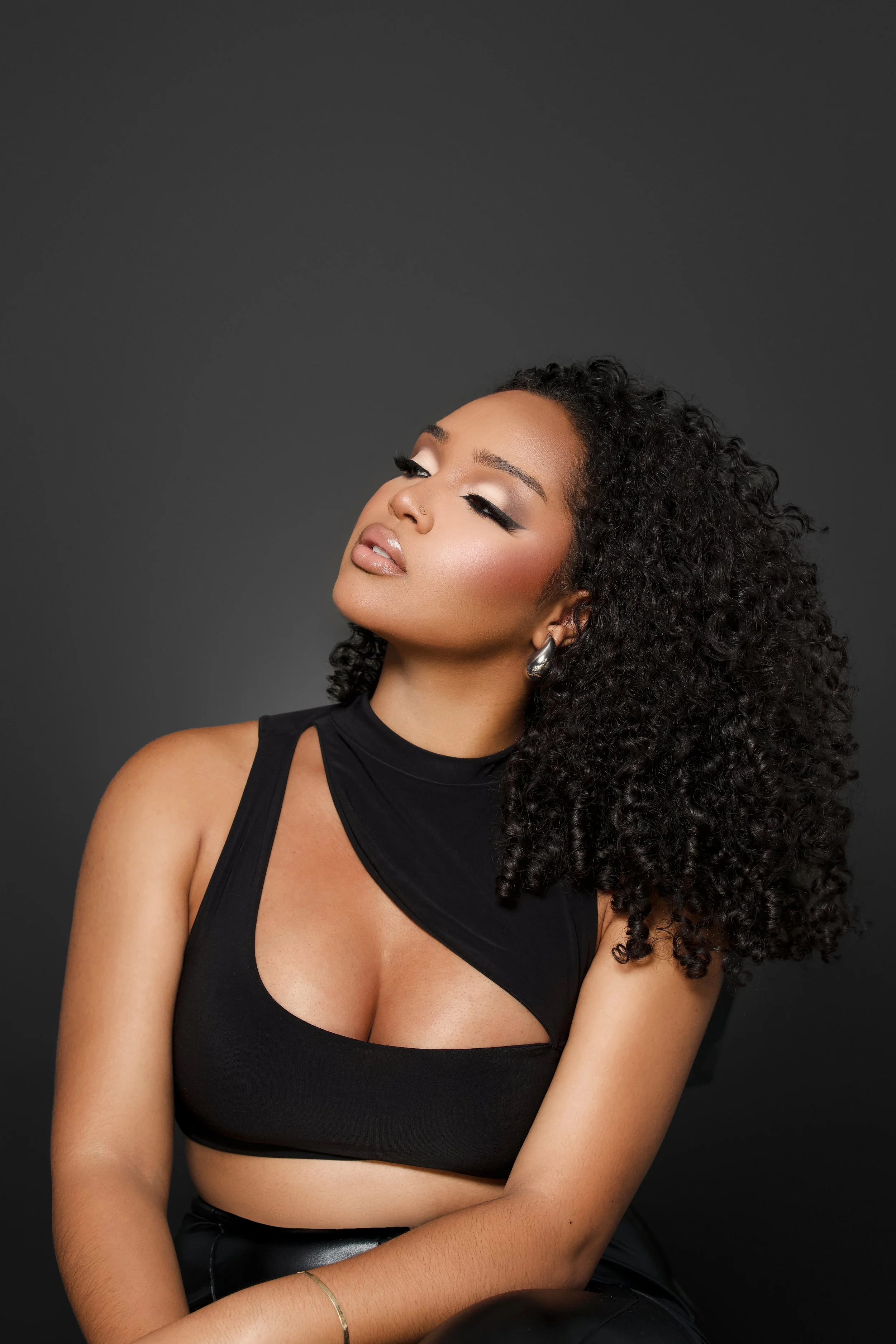 A young woman with curly black hair, wearing a black sleeveless top with a cut-out design, posing against a dark gray background with her eyes closed and head slightly tilted.