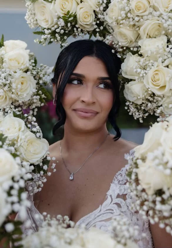 Bride with dark hair styled in loose waves, wearing a lace wedding dress and a necklace, surrounded by a floral arch of white roses and baby's breath.