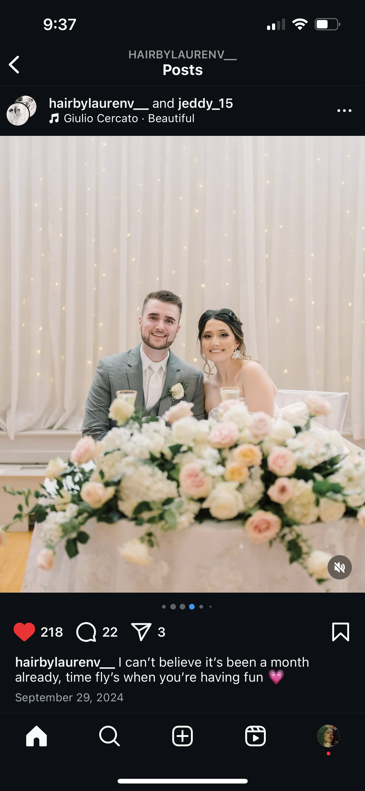 A bride and groom sitting at a wedding reception table decorated with a large floral arrangement, smiling at the camera with a backdrop of white curtains and string lights.