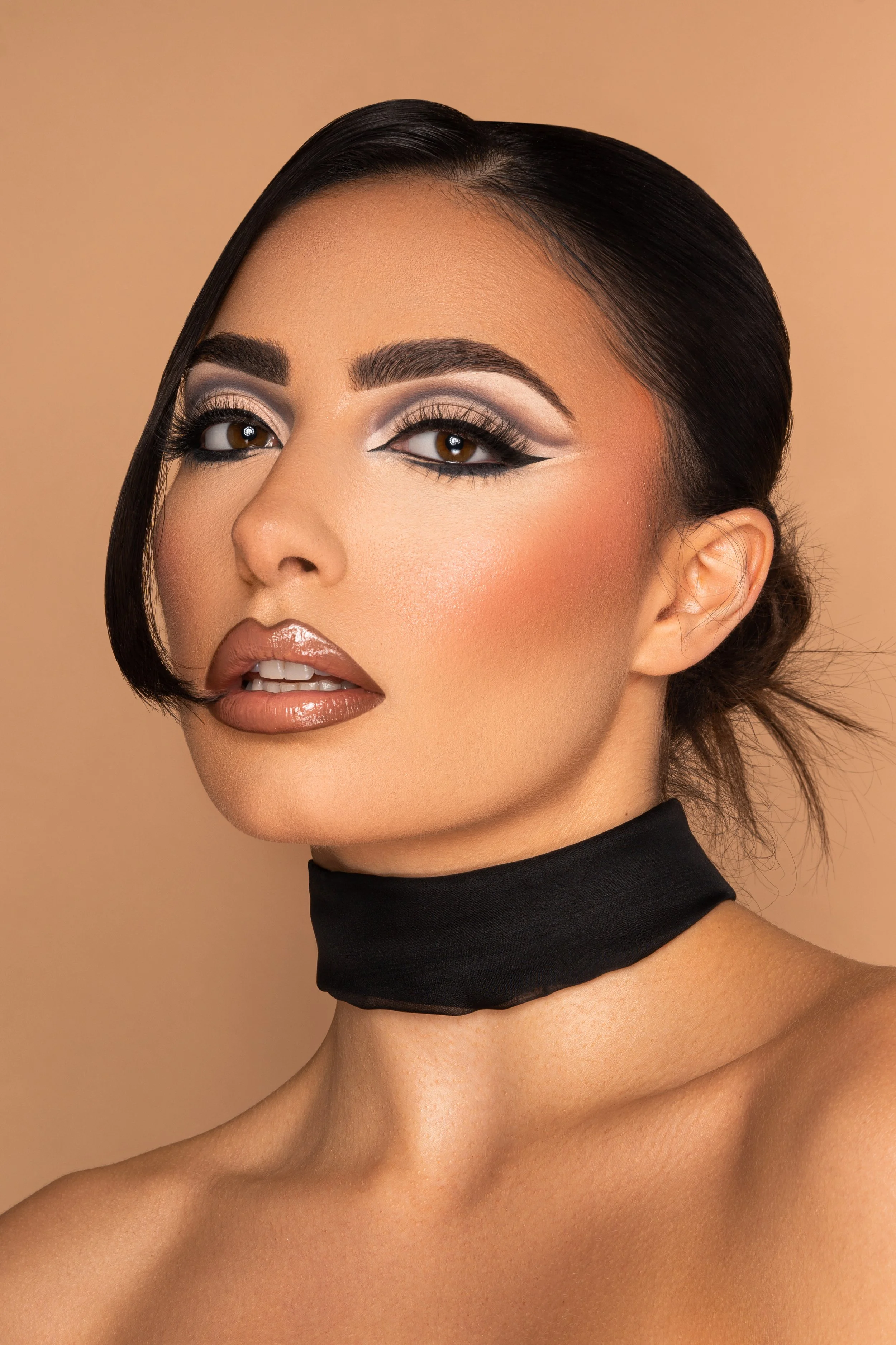 Close-up portrait of a woman with dark hair, makeup, and wearing a black choker, against a beige background.