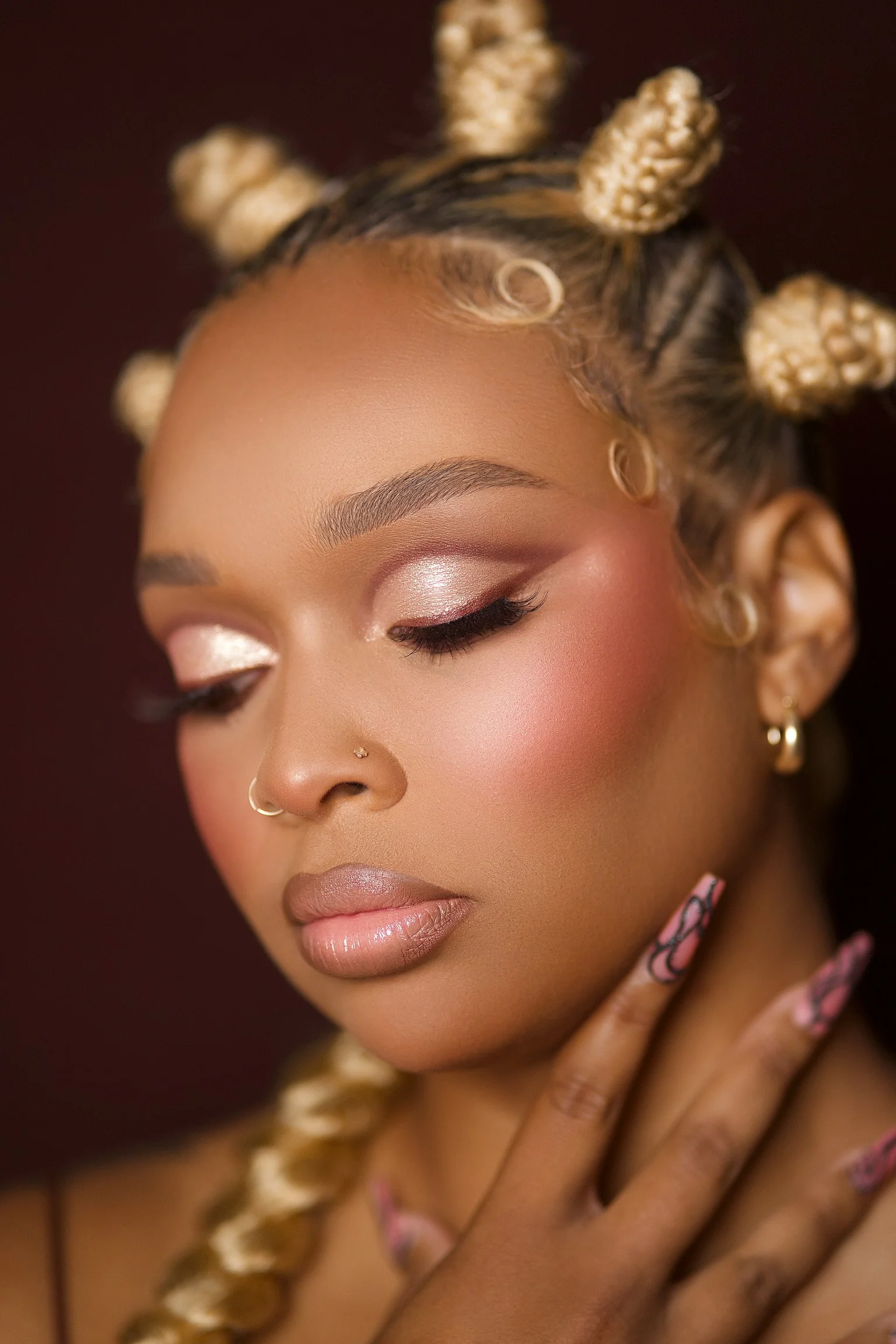 Close-up of a woman with styled blonde and black braids, wearing makeup with shimmery eyeshadow, nude lipstick, and gold jewelry, touching her face gently.