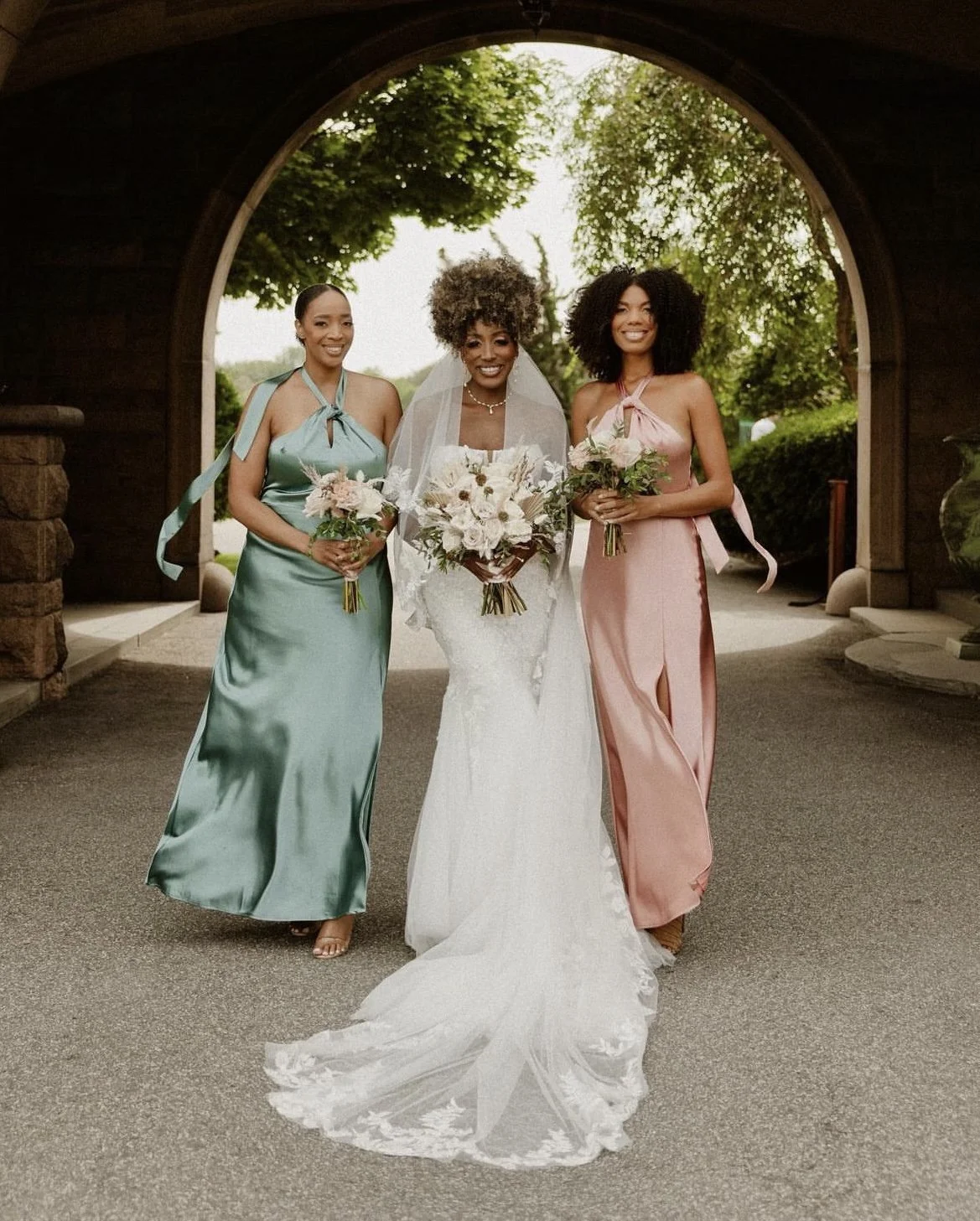 Three women, including a bride in a white wedding dress, standing under an arched wooden structure outdoors with greenery in the background. The bride holds a bouquet, and the other two women, possibly bridesmaids, hold smaller bouquets.