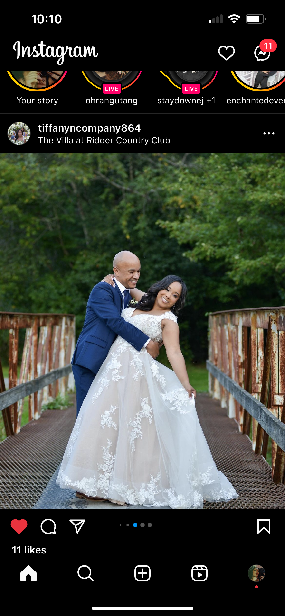 A joyful couple in wedding attire standing on an old rusty bridge outdoors, with the groom lifting the bride. The bride wears a white wedding gown with floral embroidery, and the groom wears a blue suit. They are smiling and embracing each other, wit