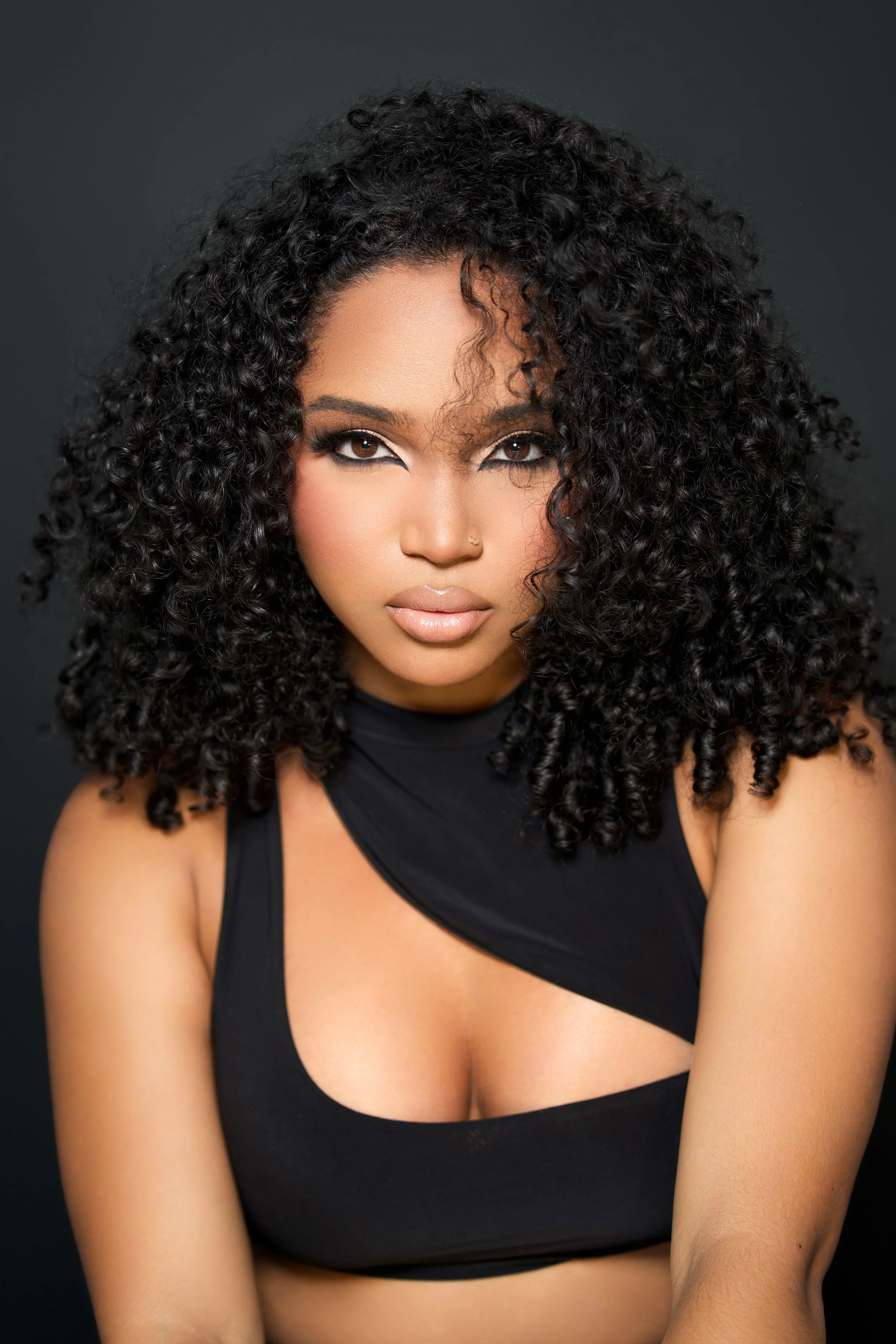 Portrait of a woman with curly black hair, makeup, and a nose piercing, wearing a black top with cutouts, against a dark background.