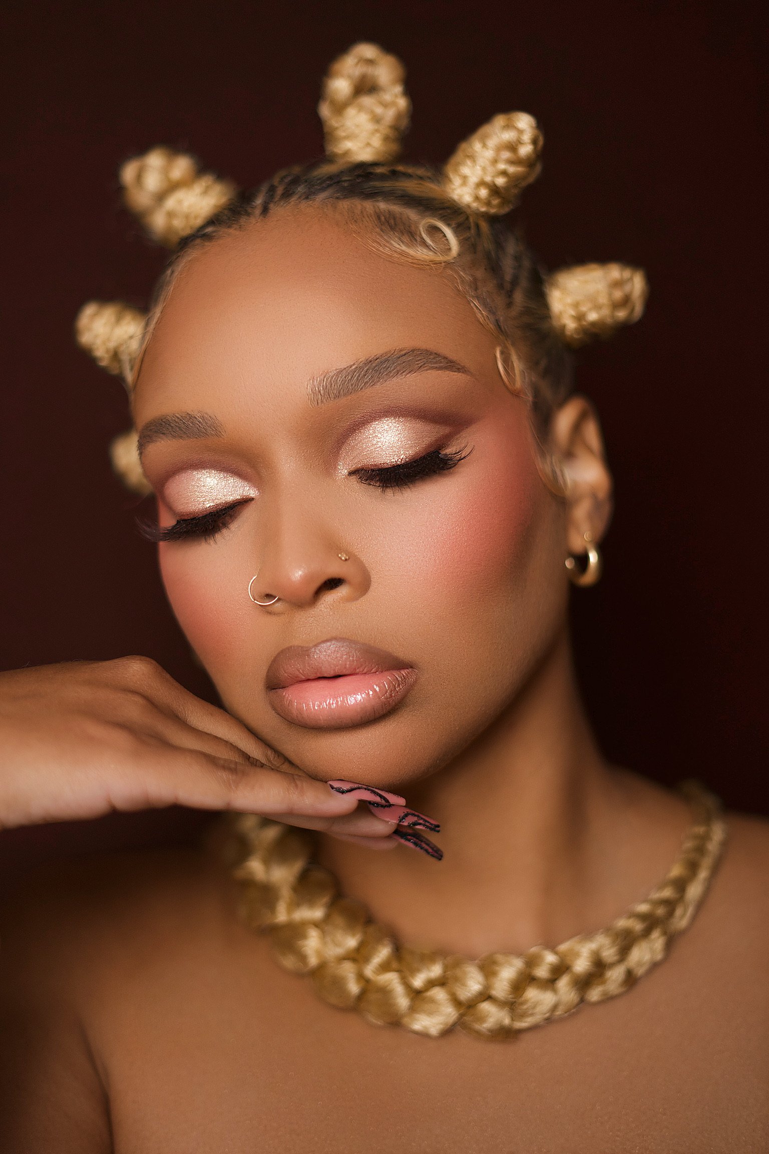 Close-up of a woman with braided blonde hair styled in bantu knots, wearing gold jewelry, with eye makeup, glossy lips, and her hand resting under her chin.