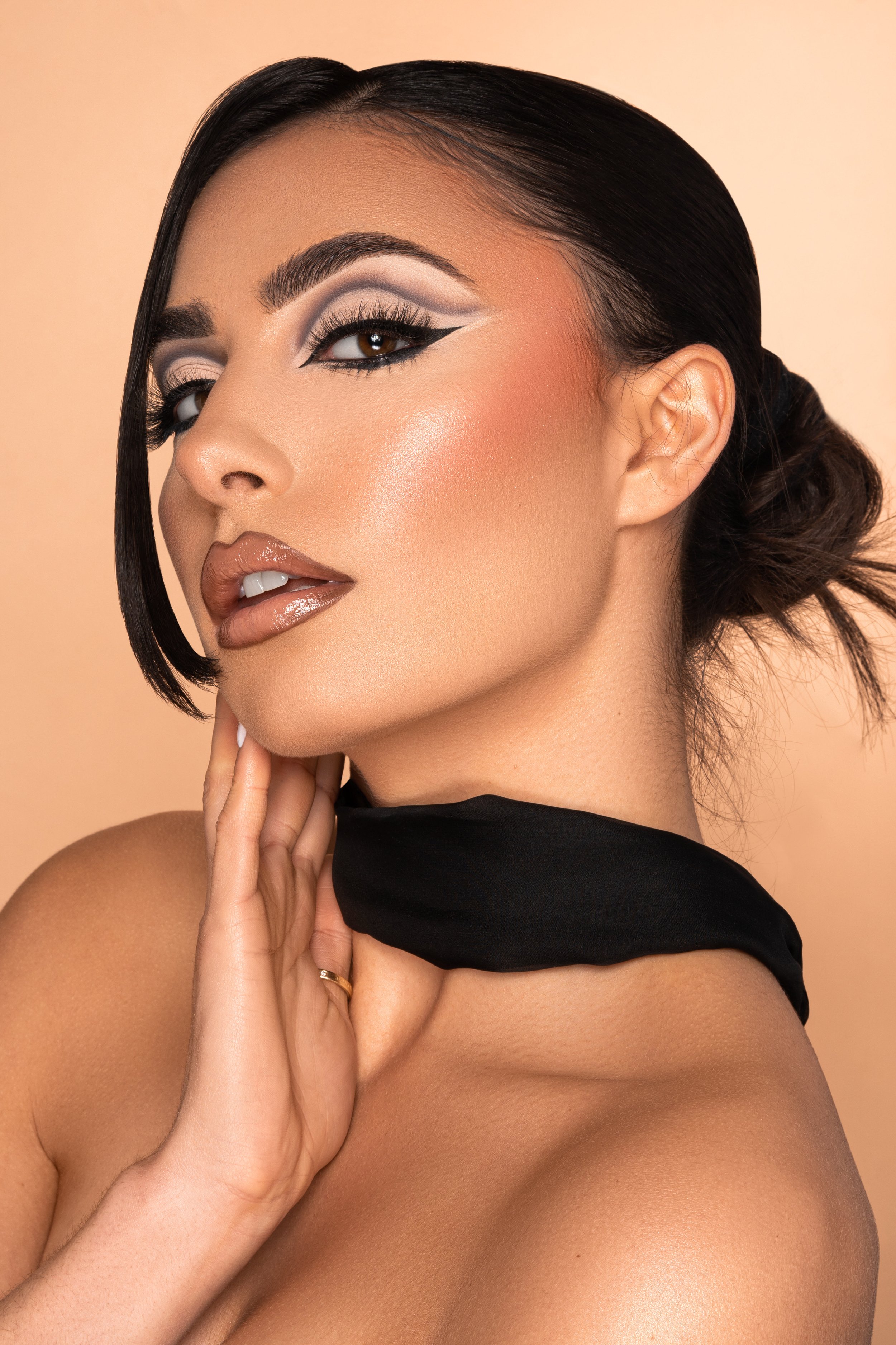 A woman with dark hair styled in a low bun, wearing bold makeup including eyeliner, eyeshadow, and nude lipstick, and a black scarf around her neck, posing against a beige background.