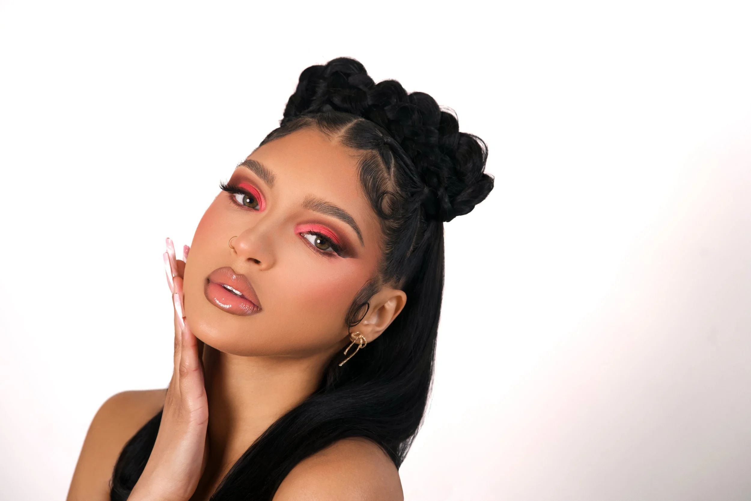 A woman with long black hair styled in intricate braids, wearing makeup with pink eyeshadow, nude lips, and hoop earrings, posing with her hand near her face against a plain background.