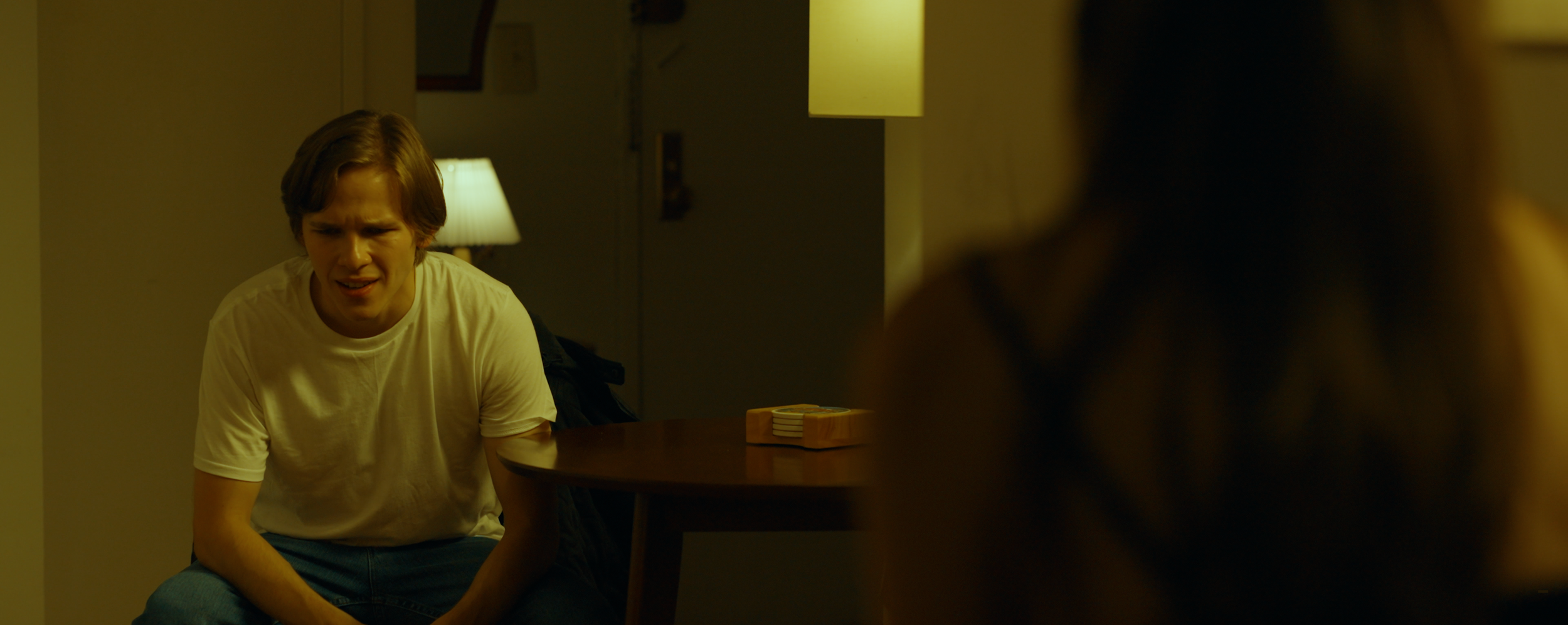A young man with light brown hair wearing a white t-shirt and blue jeans is sitting at a table, appearing distressed or confused, in a dimly lit room with warm lighting.
