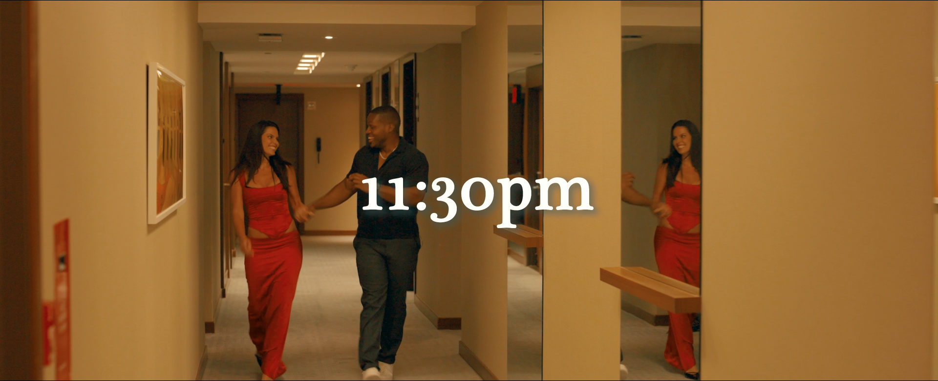 A couple walking down a hotel hallway, smiling and holding hands, with a large mirror reflecting their image, overlaid with the time 11:30 pm.