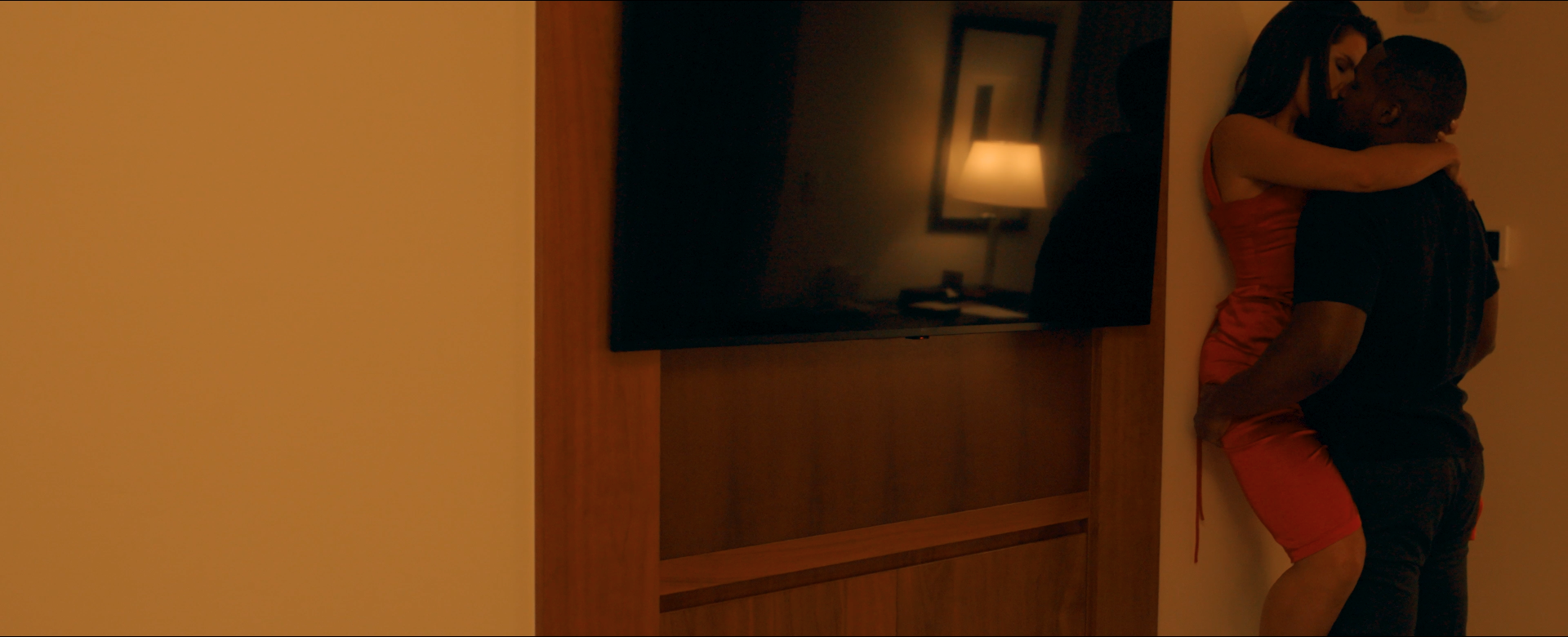 A man and woman embrace and kiss in a warmly lit hotel room, with a wall-mounted TV and lamp reflected in a mirror in the background.