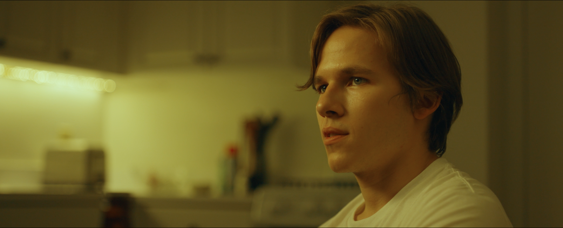 Close-up of a young man with brown hair and blue eyes, looking thoughtful, in a dimly lit room with yellowish lighting.
