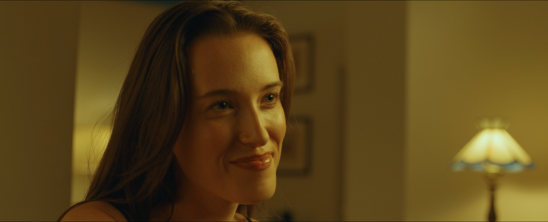 Close-up of a smiling woman with long brown hair in a warmly lit room, with a lamp in the background.