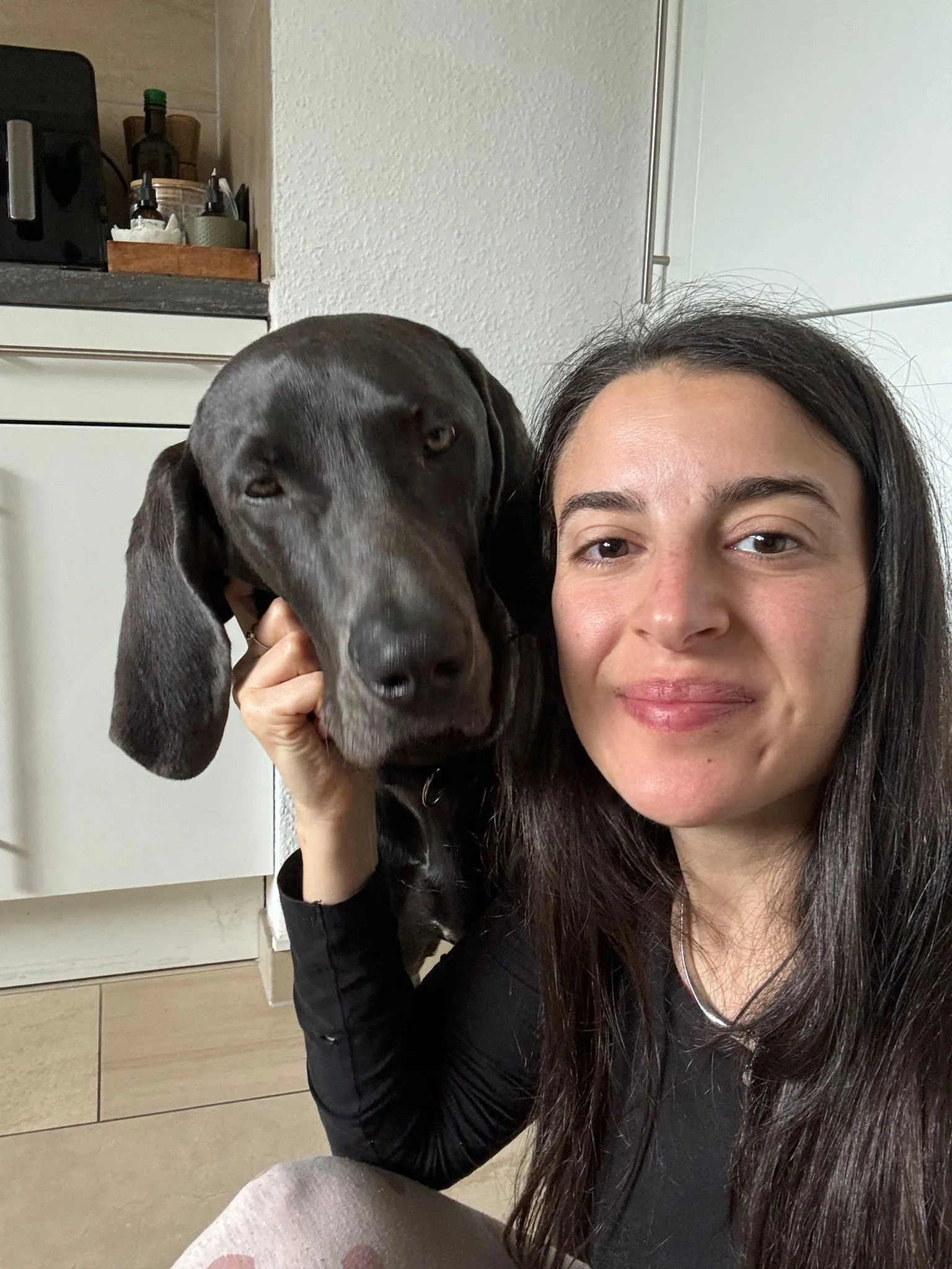 A woman with long dark hair and a black top smiling in a kitchen next to a dark gray dog with long ears, who is resting its head on her shoulder.