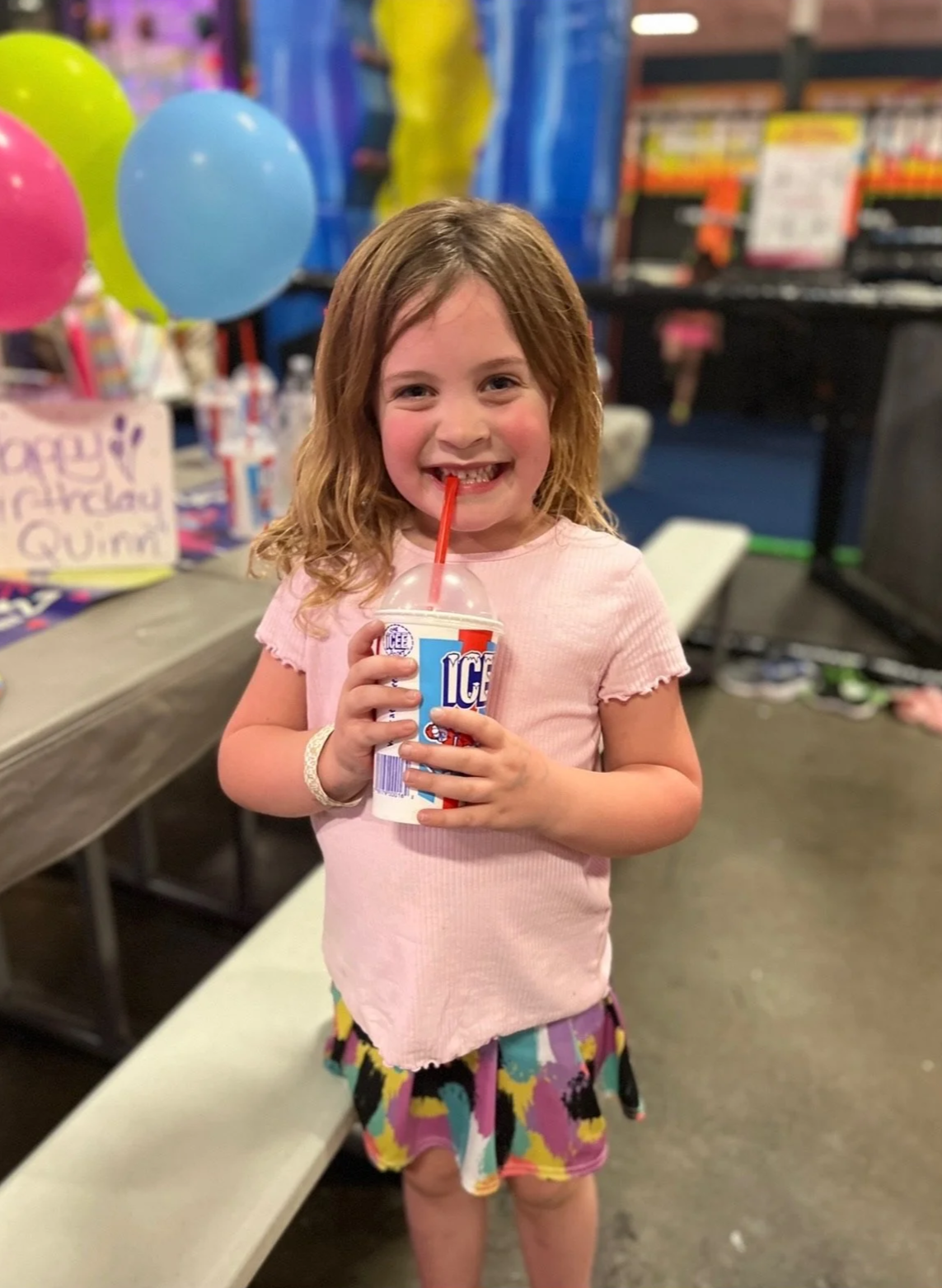 A young girl with shoulder-length brown hair enjoys a McDonald's ice cream sundae with a red straw. She is smiling widely, showing her teeth, and wearing a light pink shirt and a colorful, pattern skirt. Behind her are balloons and a birthday party s