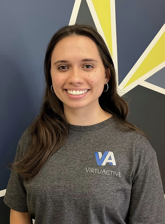 Young woman with long brown hair wearing a gray T-shirt with 'VA VirtuActive' logo, smiling, standing in front of a geometric wall pattern.