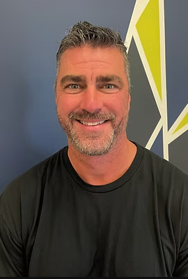 A smiling man with short, gray hair and a beard, wearing a black shirt, standing in front of a colorful geometric background.