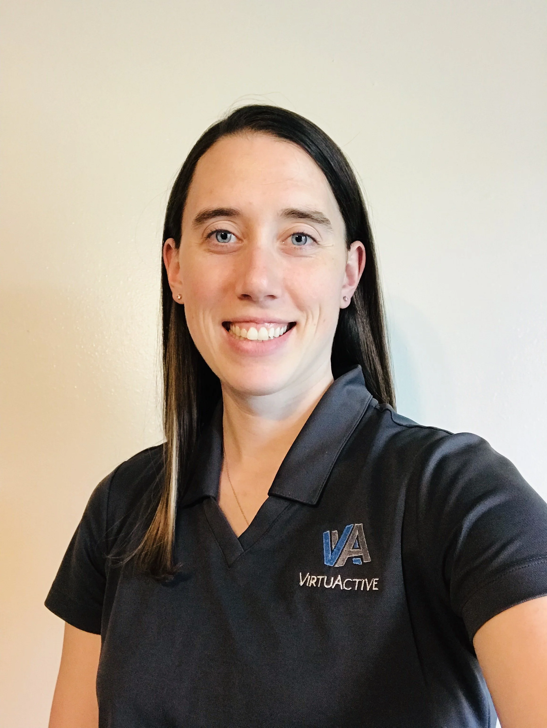 A woman with long dark hair smiling, wearing a black collared shirt with a "VIRTUAACTIVE" logo on it, standing in front of a plain light-colored wall.