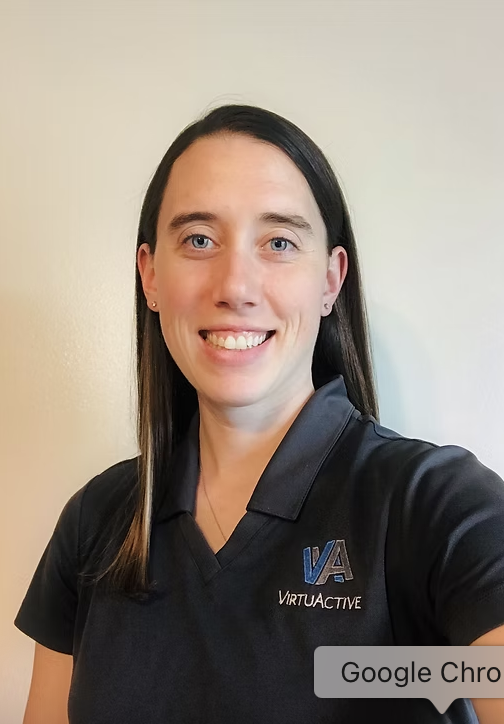 A woman with long dark hair smiling, wearing a black collared shirt with a "VIRTUAACTIVE" logo on it, standing in front of a plain light-colored wall.