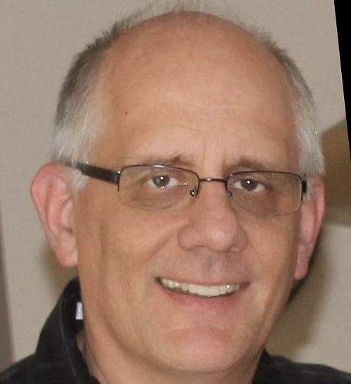 A smiling man with short, gray hair and a beard, wearing a black shirt, standing in front of a colorful geometric background.