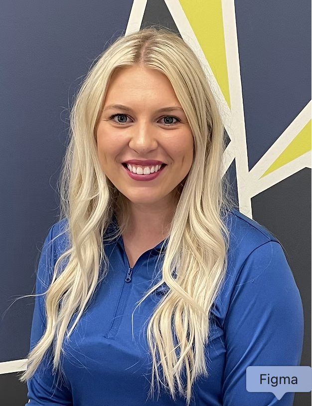 A woman with long blonde hair wearing a blue zip-up shirt, smiling at the camera, standing in front of a colorful geometric wall design.