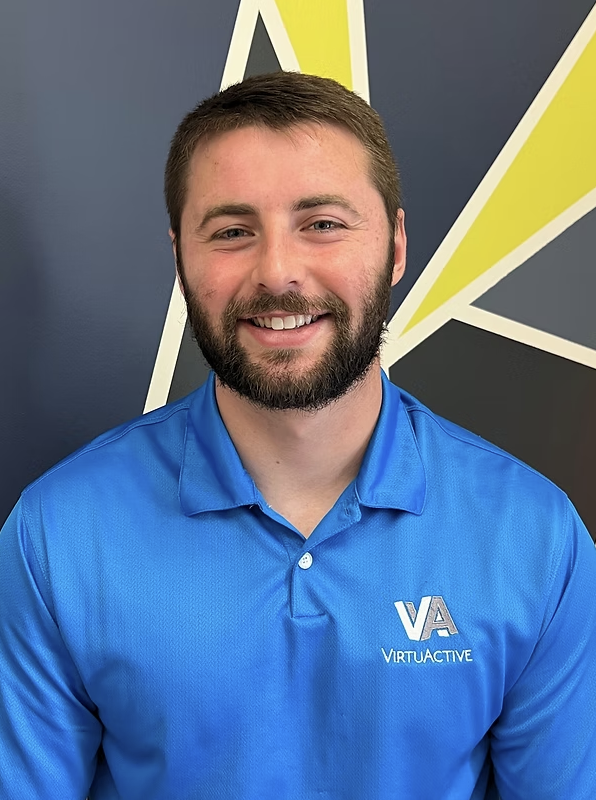 A smiling man with a beard and short brown hair, wearing a blue shirt with the VU VirtuActive logo, standing in front of a dark wall with yellow, white, and gray geometric designs.