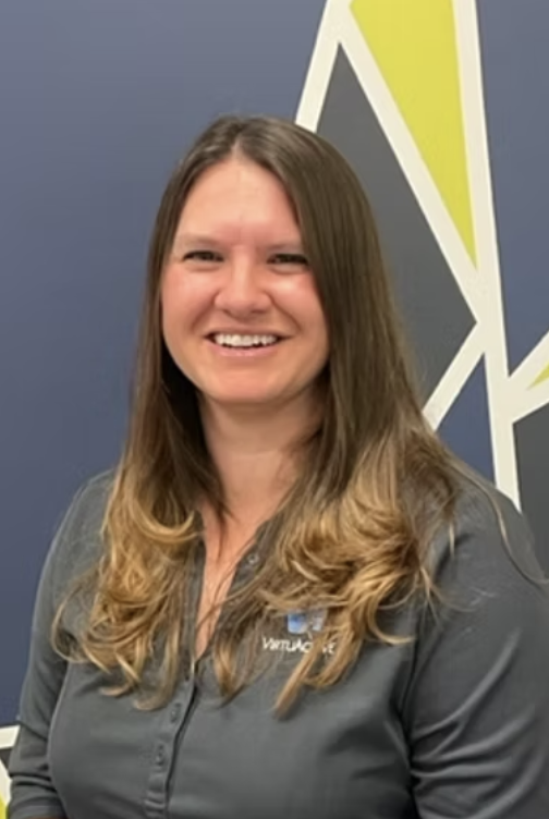 woman with long brown hair smiling, wearing a gray jacket with a logo, standing in front of a blue wall with a geometric design.